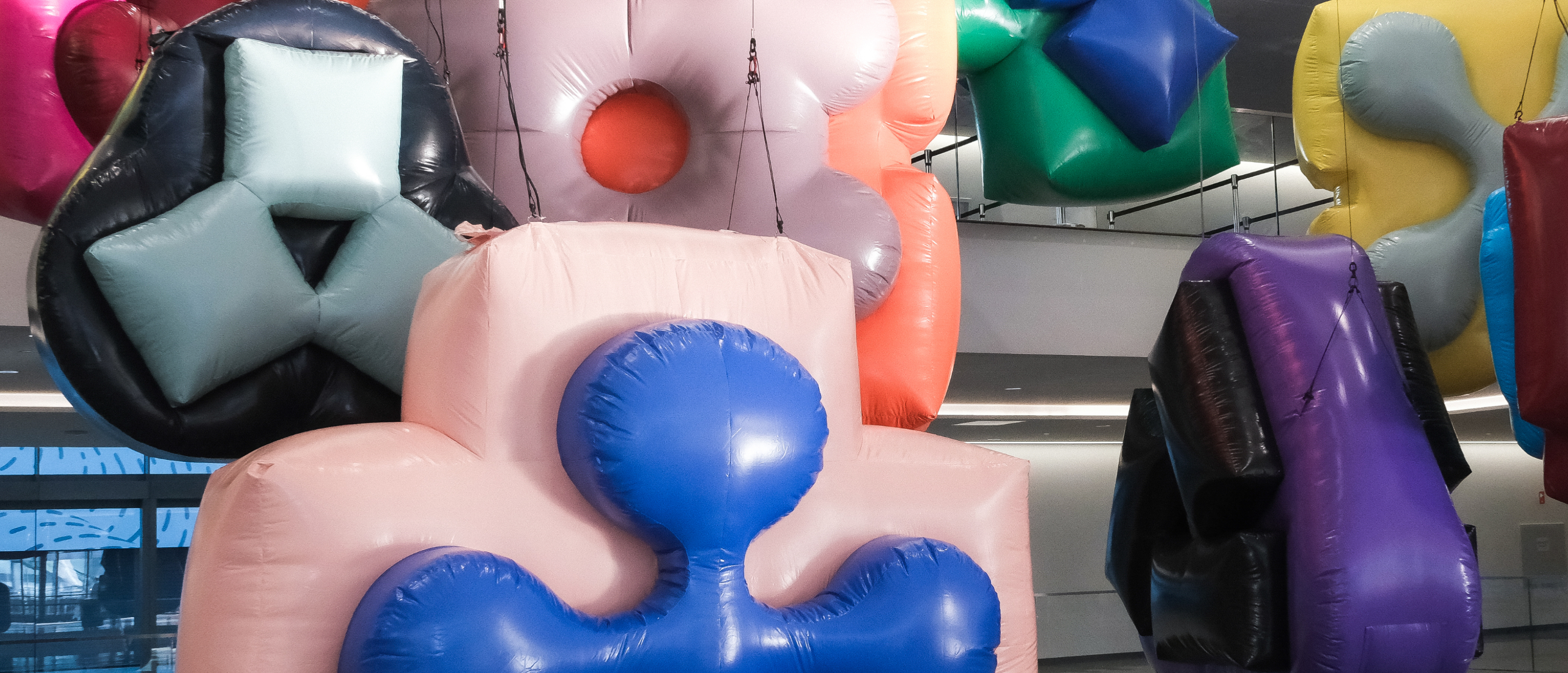 Inflatable sculptures in various abstract shapes and colors hang from a ceiling in bright indoor light