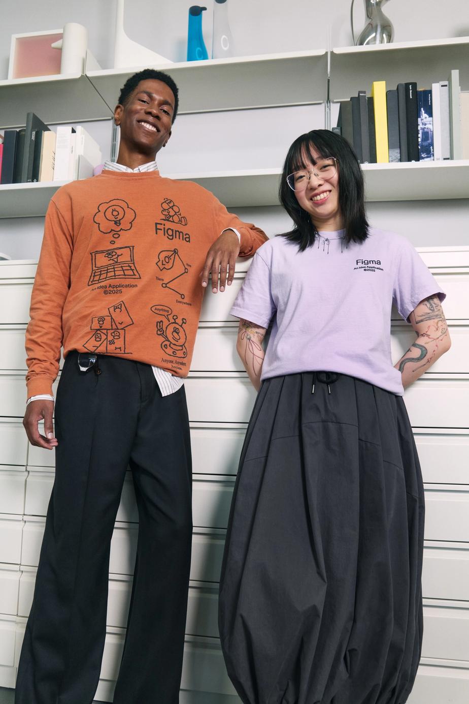 Two people pose in front of office shelves wearing Figma shirts—one in an orange long-sleeve, the other in a lavender tee, smiling confidently.