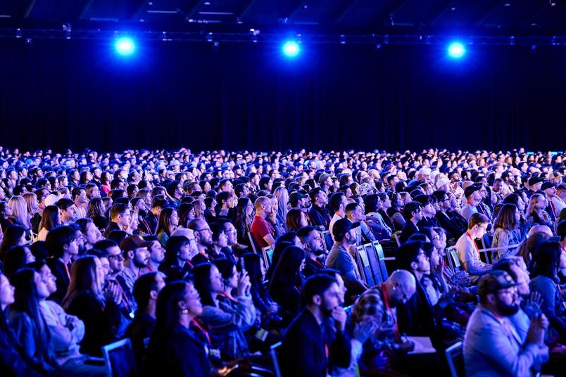 A large seated audience in an auditorium, illuminated by blue overhead lights, attentively focused on an unseen stage.