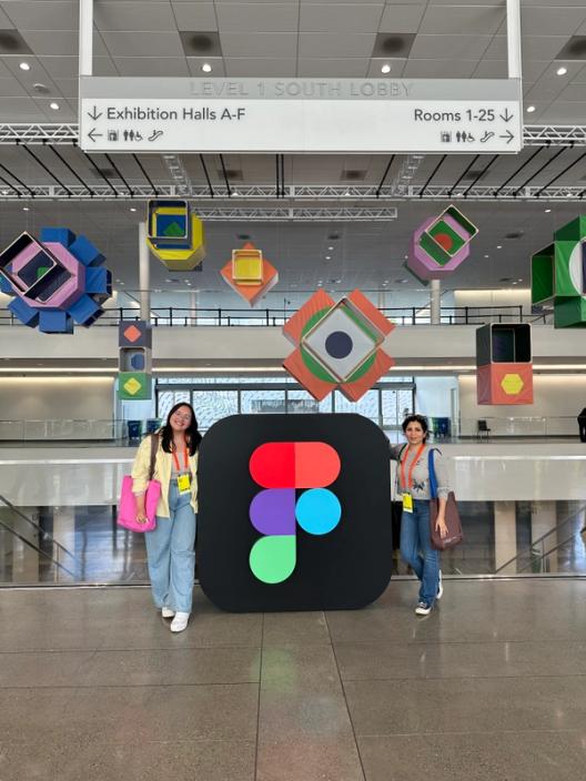 Two women pose on either side of a large Figma logo, with supergraphic decorations hanging from the ceiling in the background.