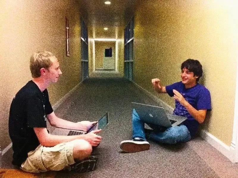 Two young men sit in a dorm hallway, chatting with laptops on their laps, smiling animatedly.