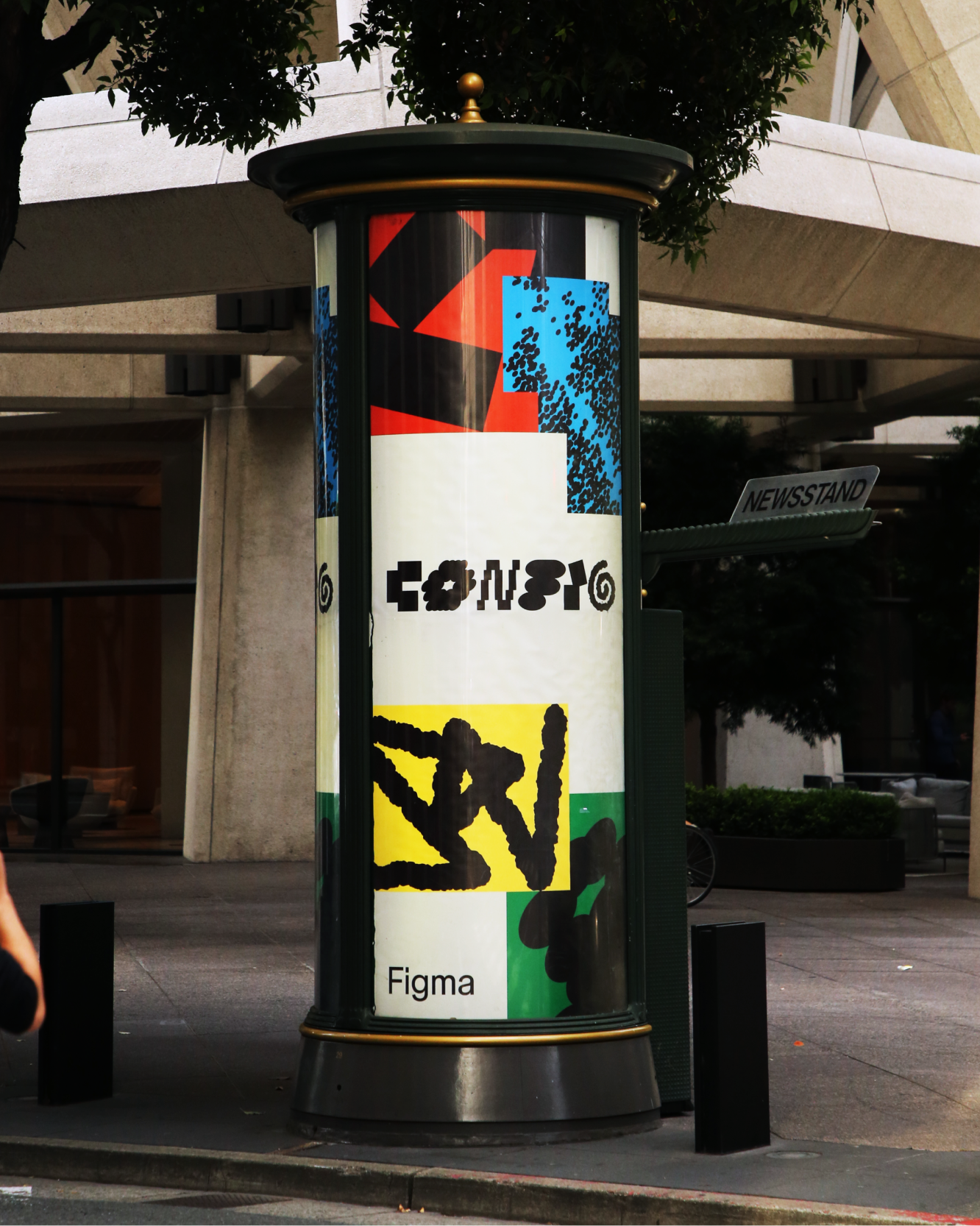 A colorful Config ad wraps a street kiosk under a tree