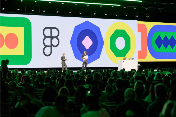 In a dark event space, there is a long stage with two people standing addressing the large audience in the foreground. Behind the presenters, there are large screens with brightly-colored shapes filling the space.