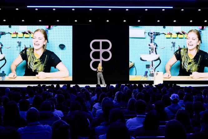 A speaker stands on stage in front of two large screens showing the same image of a woman laughing with a messy workbench in the background. The Figma logo is displayed on the screen behind them.