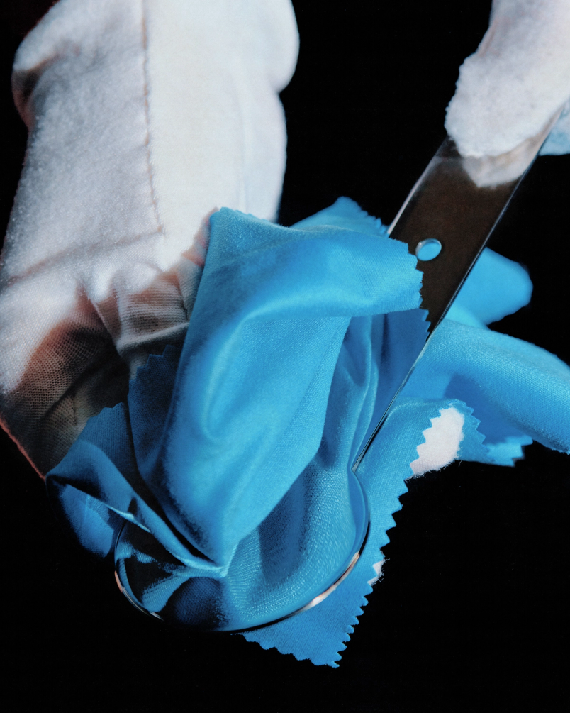 Gloved hands polish a shiny metal utensil using bright blue cloth against a dark background.