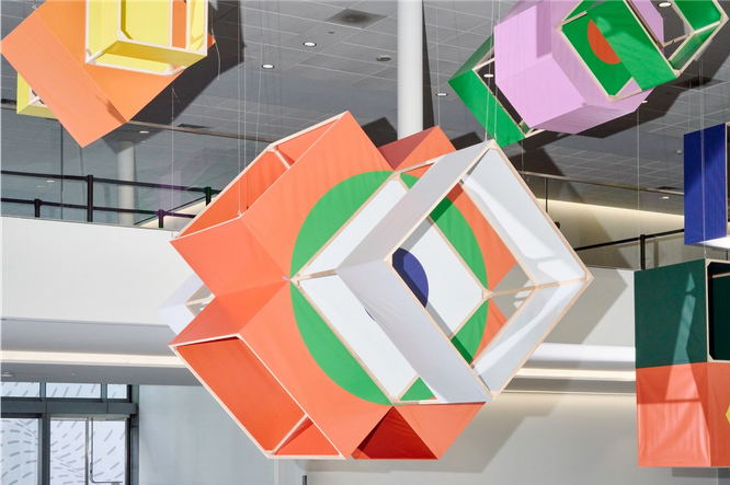 Four box kites are hanging from ceiling rafters over a large conference space. Each kite is a different 3D shape, with diamonds, squares, octagons, and circles. Some panels of the box kites are filled with orange, green, navy, yellow, or grey fabric, and some panels are empty.
