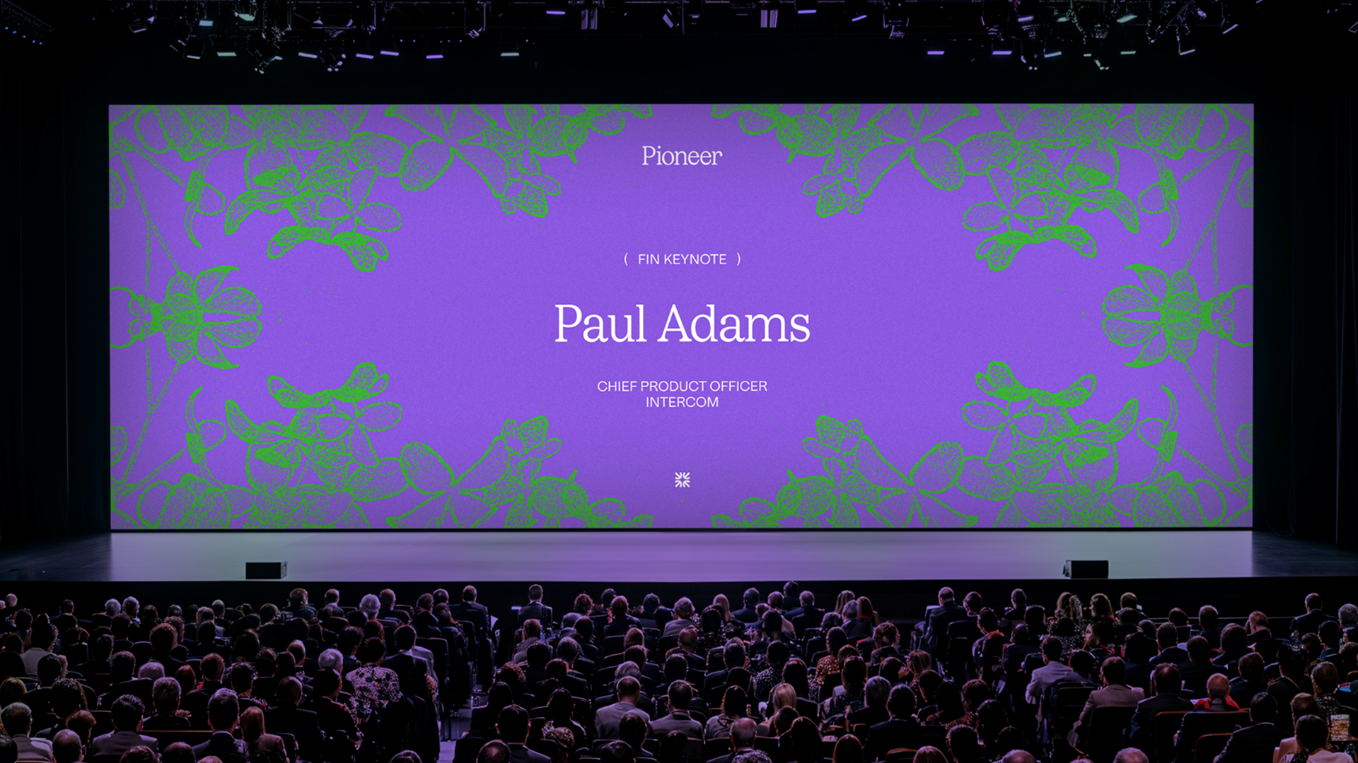 A dark presentation room with a large purple screen and green flowers announces speaker Paul Adams.