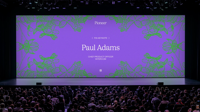 A dark presentation room with a large purple screen and green flowers announces speaker Paul Adams.