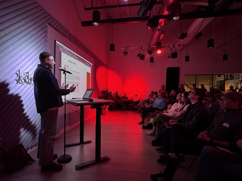 Dark room with a full audience watching speaker with red hued presentation