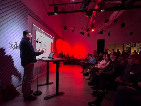Dark room with a full audience watching speaker with red hued presentation