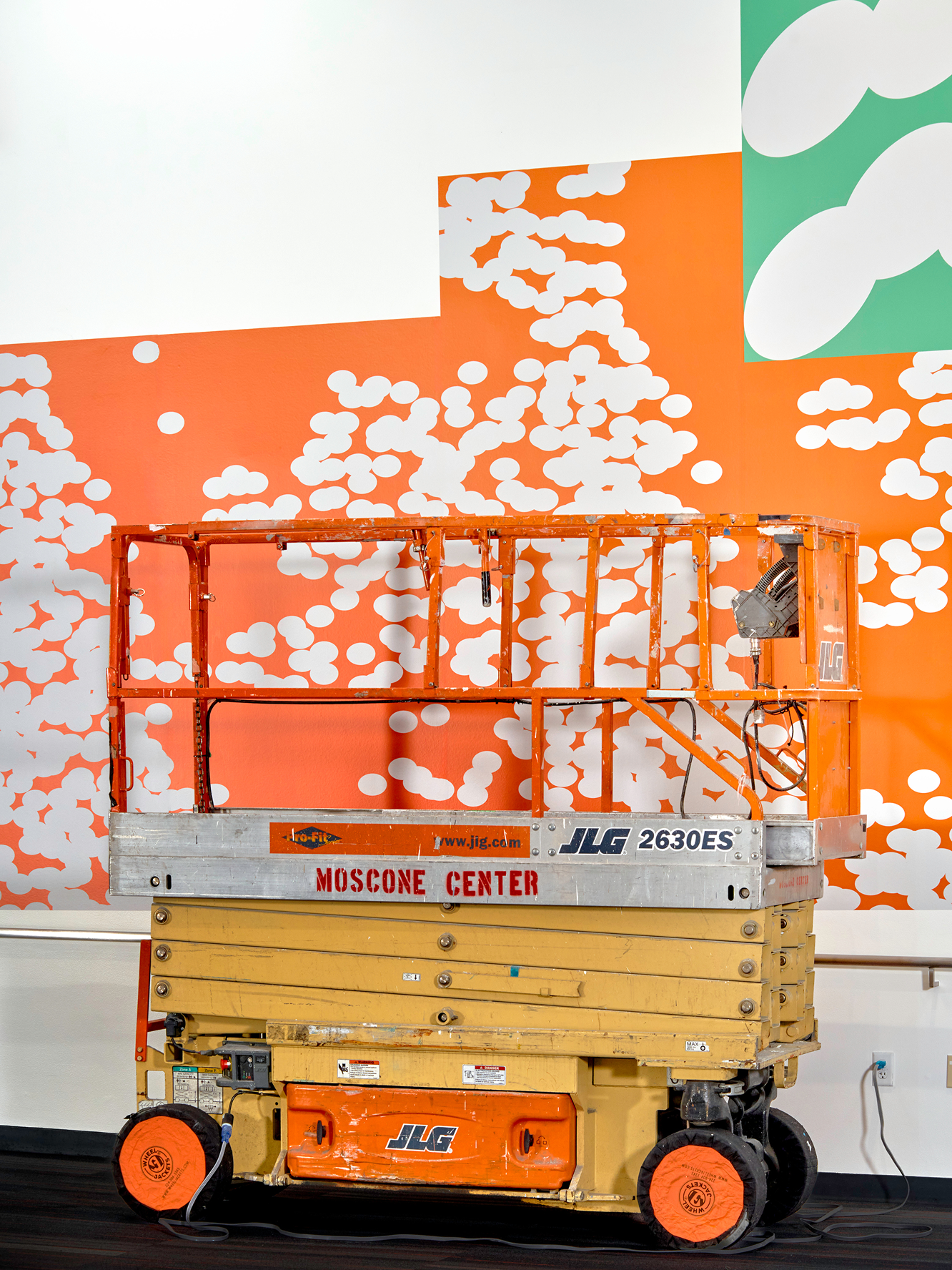 A scissor lift sits in front of an orange and green dot-patterned mural
