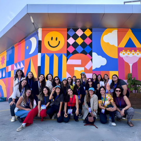 A group of women posing outdoors in front of a colorful mural with geometric patterns, smiley faces, and other bright designs. They are smiling, wearing event badges and casual attire.