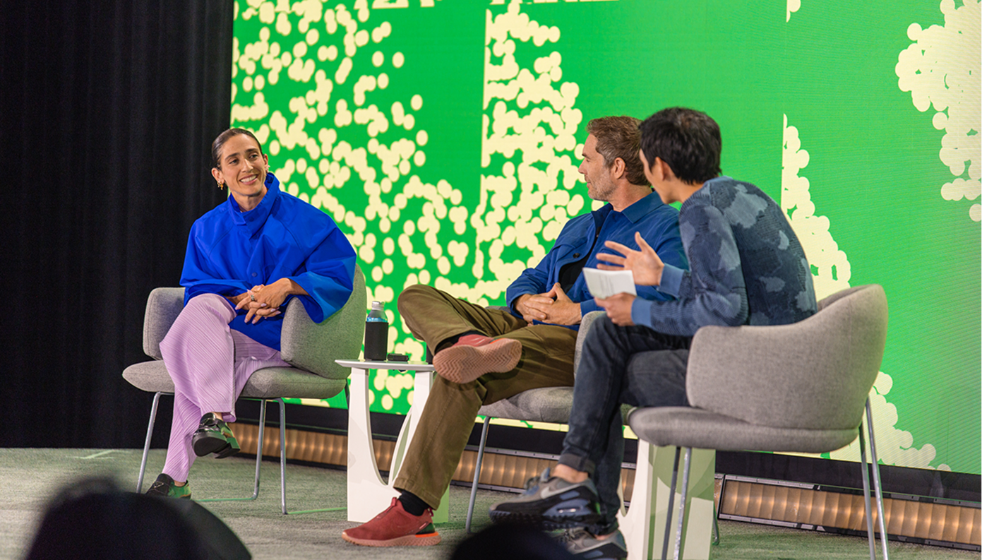 Rachel Been, Charlie Sutton, and Yuhki Yamashita sit in gray armchairs on a stage in front of a bright green patterned screen, mid-conversation.