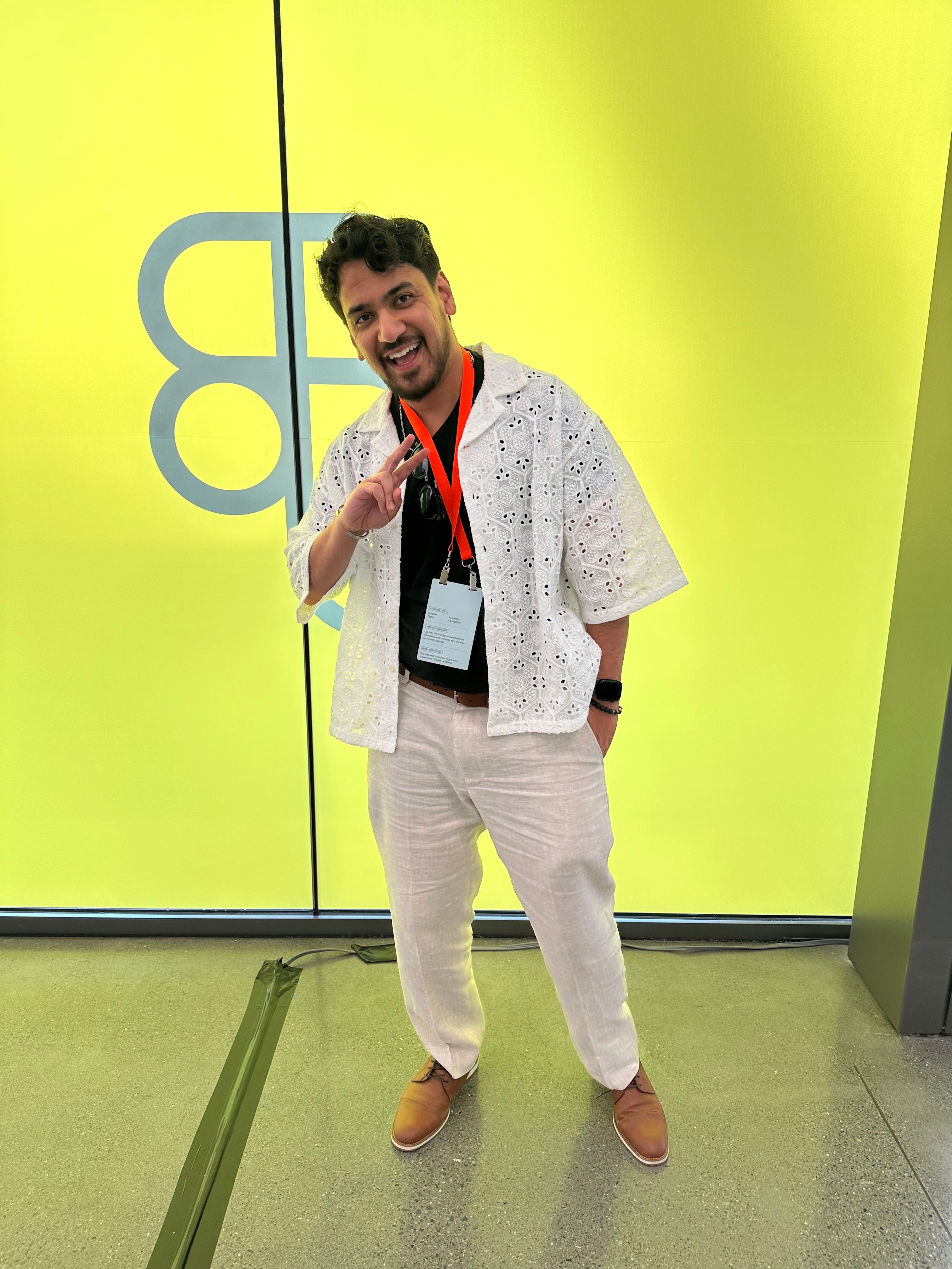 A man wears an open white lace shirt over a black tee and white pants