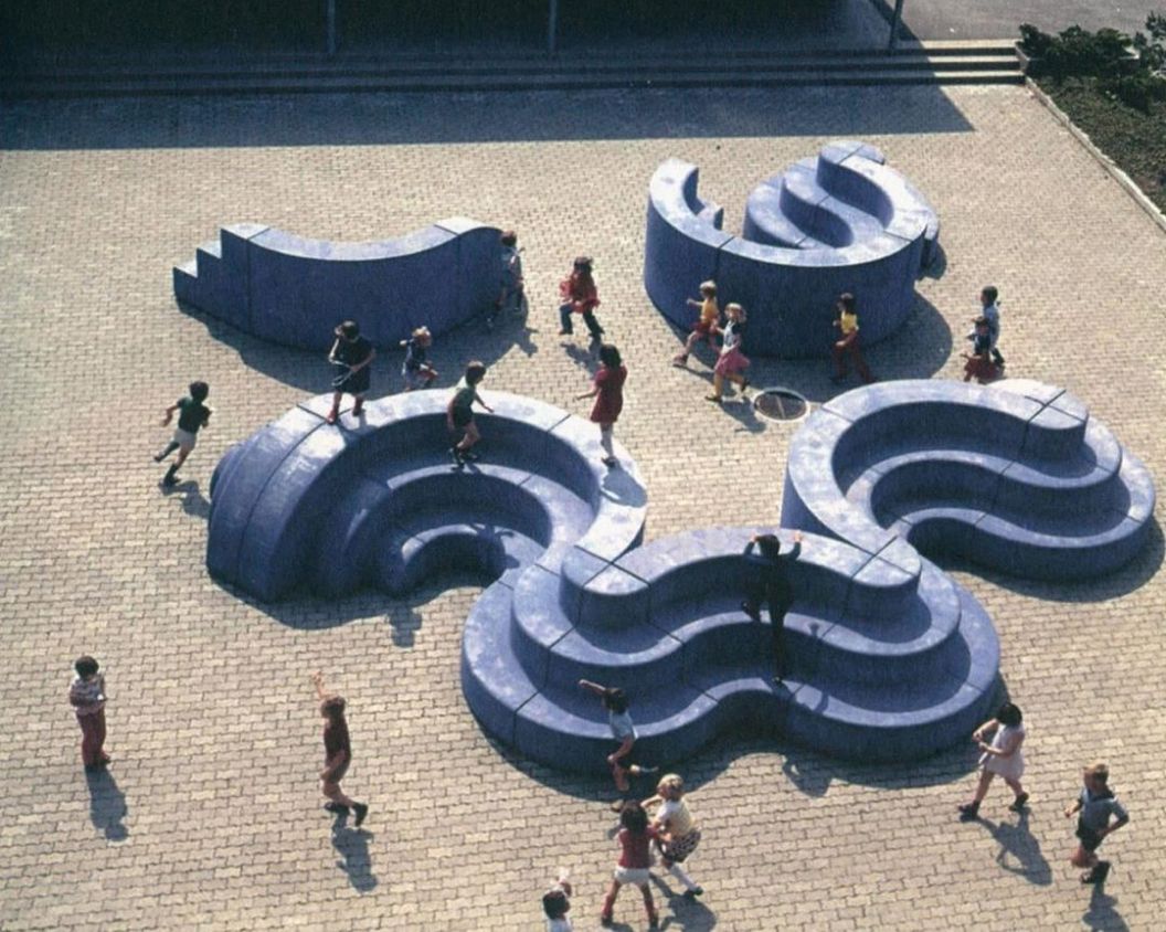 A playground filled with children playing on large, blue, wavy, and circular structures. The play structures resemble flowing, multi-level curves, creating spaces for climbing, sitting, and running. The ground is paved with tiles, and children are seen interacting with the dynamic shapes.