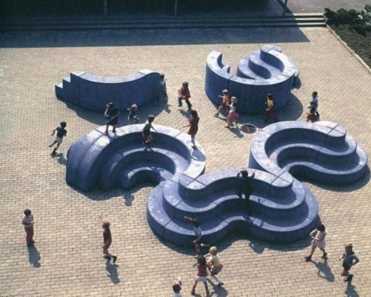 A playground filled with children playing on large, blue, wavy, and circular structures. The play structures resemble flowing, multi-level curves, creating spaces for climbing, sitting, and running. The ground is paved with tiles, and children are seen interacting with the dynamic shapes.