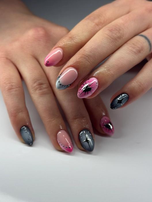 A close-up photograph of a pink and gray manicure featuring almond-shaped nails with small gems, starburst patterns, and metallic accents.