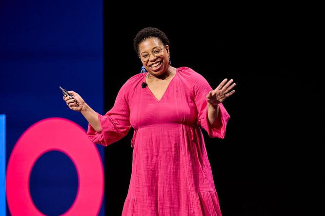 A speaker in a bright pink dress smiles and gestures enthusiastically while presenting on stage. The background features a partial view of large, colorful letters on a dark screen.
