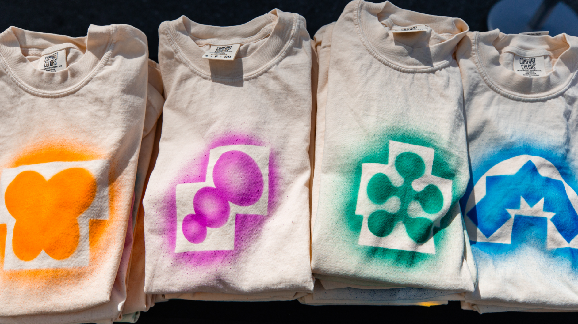 Four folded white t-shirts display spray-painted Config icon designs in orange, purple, green, and blue.