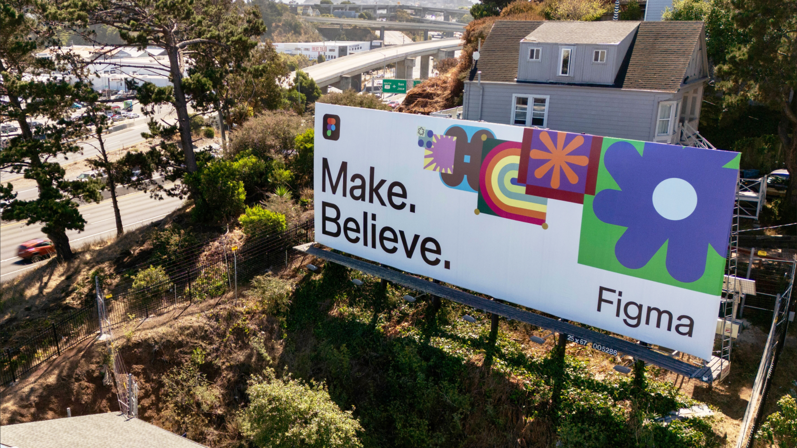 A horizontal Figma billboard positioned along a highway near a hillside covered in trees. The sign displays bold text that reads "Make. Believe." on a white background, with colorful abstract icons, including a large purple flower, a starburst shape, and a rainbow-like pattern. The Figma logo appears on the left, and "Figma" is written in black text on the right side of the billboard. A residential house and distant roads are visible in the background.