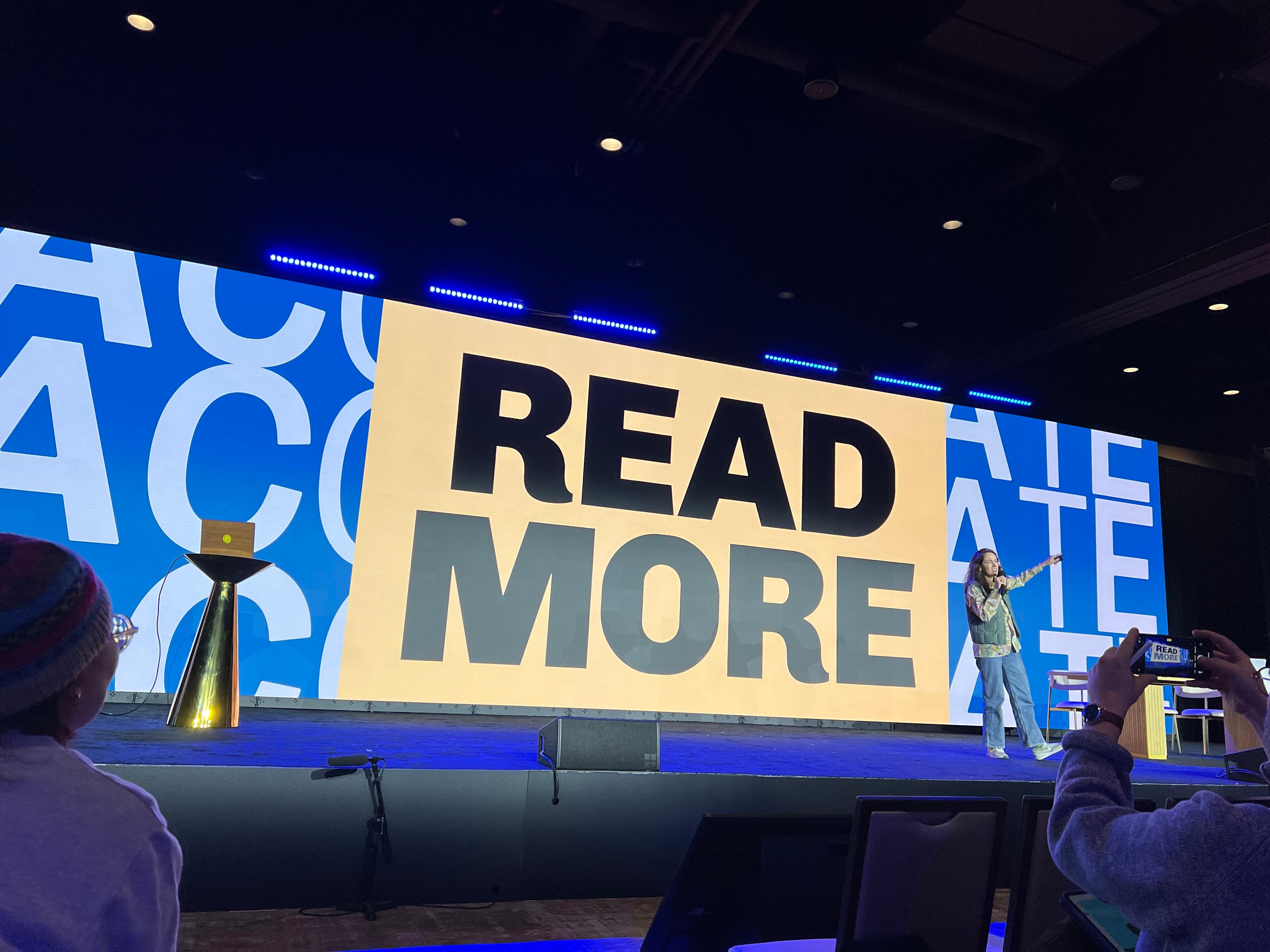 Our author commands the stage with confidence, gesturing towards a massive screen that boldly declares "READ MORE" in striking black capital letters on a beige background. Dressed in a vibrant, effortlessly cool outfit, she captivates the audience with her energy and presence. The stage design is sleek and modern, illuminated by blue LED lighting, with a dynamic backdrop featuring large, partially visible white text on a blue background. In the foreground, engaged audience members take in the moment, one capturing Carly’s wisdom on their phone, while another watches attentively from beneath a colorful beanie. A gold-colored lectern with a small box on top sits to the left, adding to the scene’s polished aesthetic.