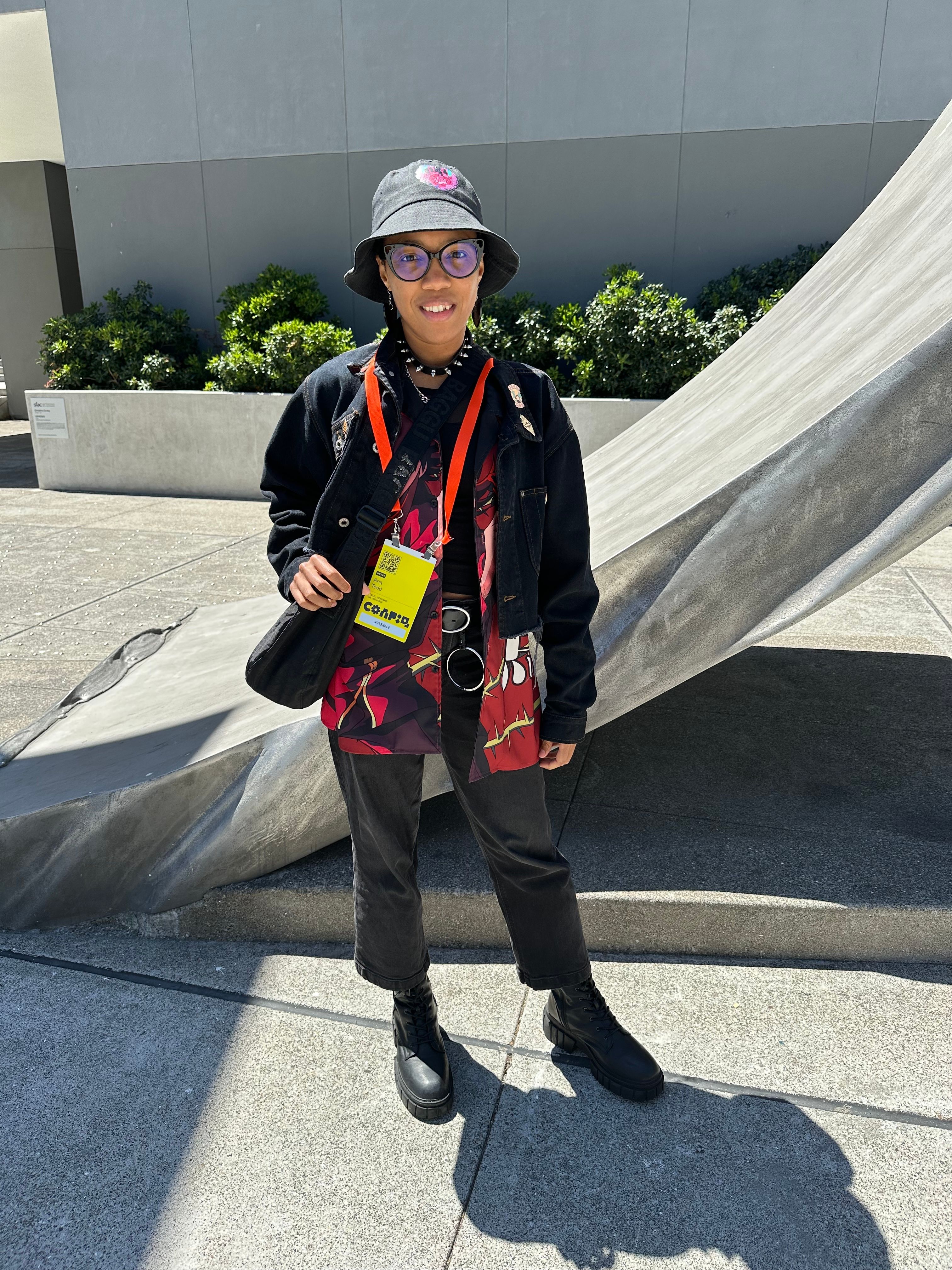 A woman wears a black bucket hat, a black jean jacket, and an anime-printed button down.