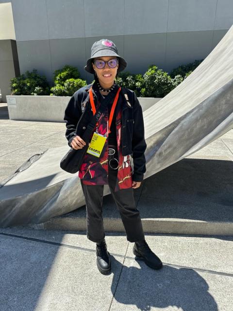 A woman wears a black bucket hat, a black jean jacket, and an anime-printed button down.