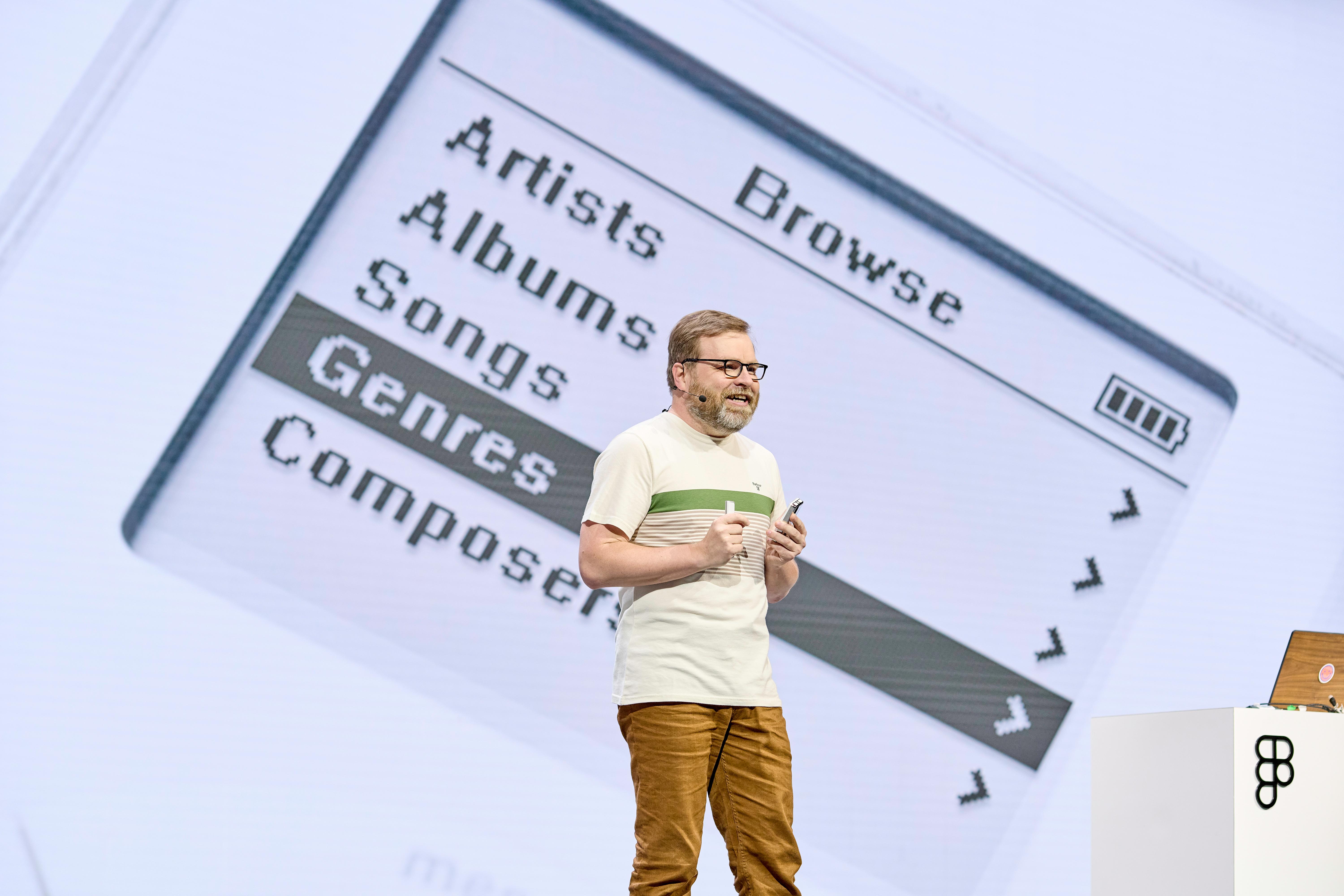 A presenter, wearing a casual white shirt with green stripes and tan pants, is enthusiastically speaking on stage. Behind him, there is a large projected screen displaying a retro-style interface of a music player with options like "Artists," "Albums," "Songs," and "Genres" highlighted.