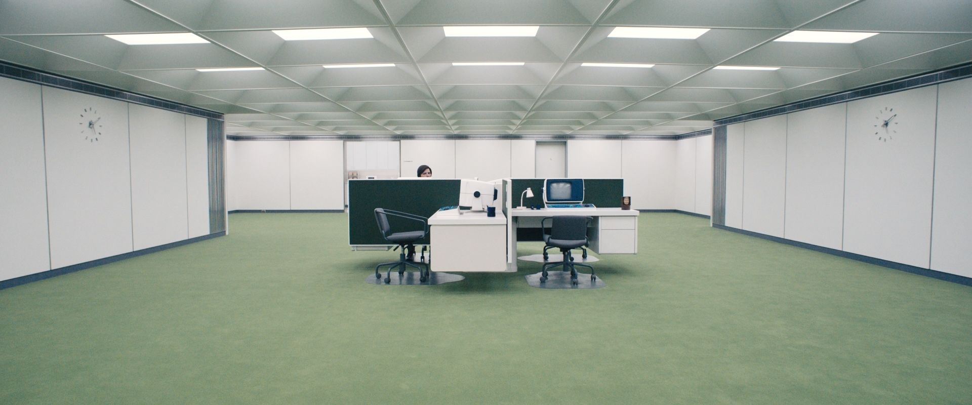 A lone worker sits at a small cluster of desks in a vast, sterile white office with green carpet and symmetrical clocks on the walls.