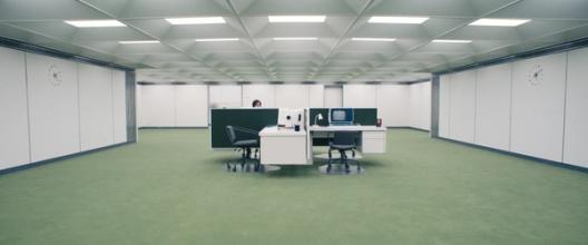 A lone worker sits at a small cluster of desks in a vast, sterile white office with green carpet and symmetrical clocks on the walls.