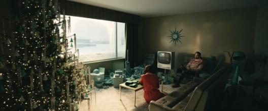 A vintage-style living room with a Christmas tree is filled with teal household items as a man and woman sit in quiet contemplation.