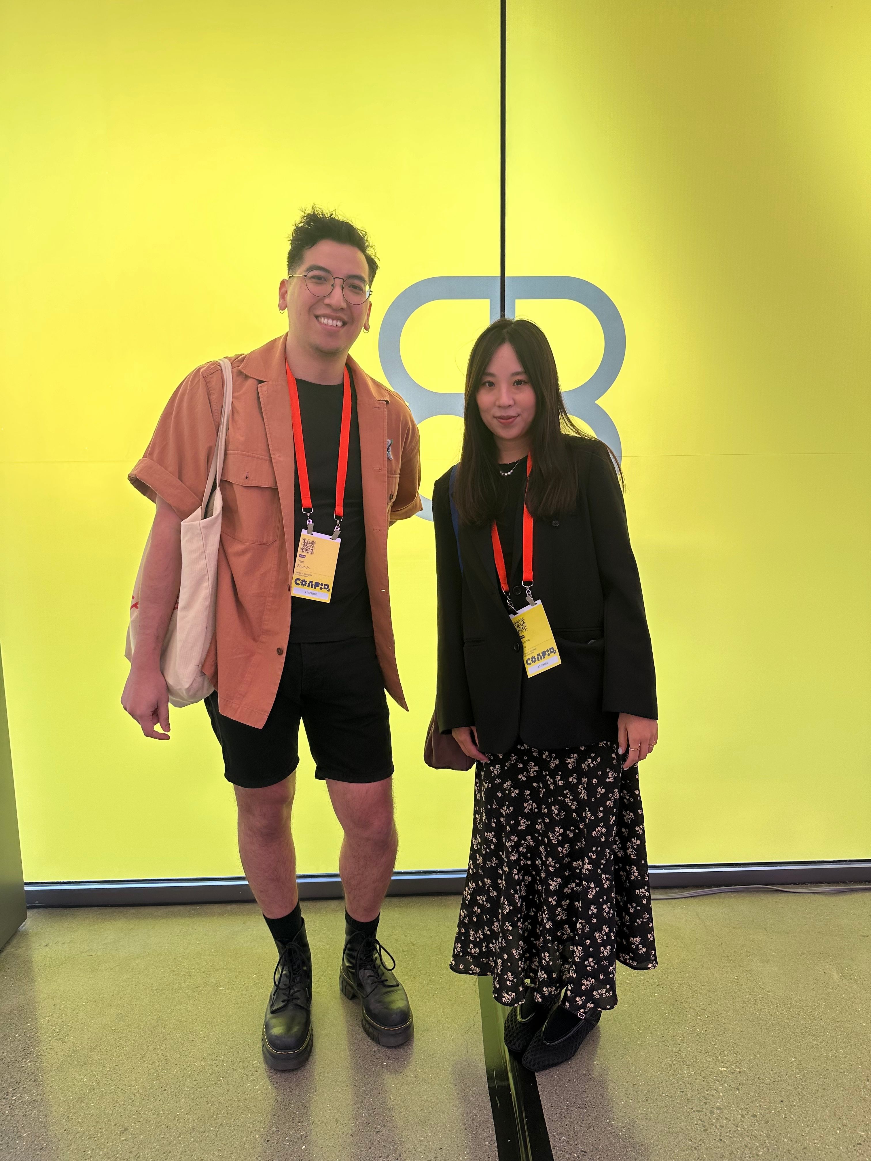 A man wears a rust orange button down over a black tee, and a woman wears a black blazer and floral maxi skirt
