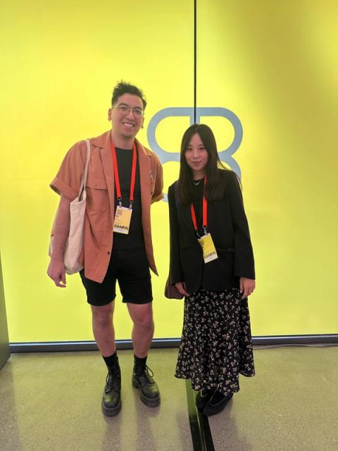 A man wears a rust orange button down over a black tee, and a woman wears a black blazer and floral maxi skirt