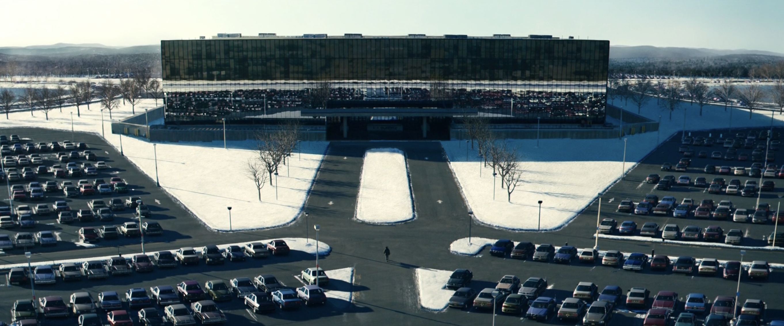 A lone figure walks across a vast parking lot toward a massive mirrored office building surrounded by snow.