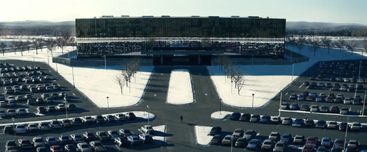 A lone figure walks across a vast parking lot toward a massive mirrored office building surrounded by snow.