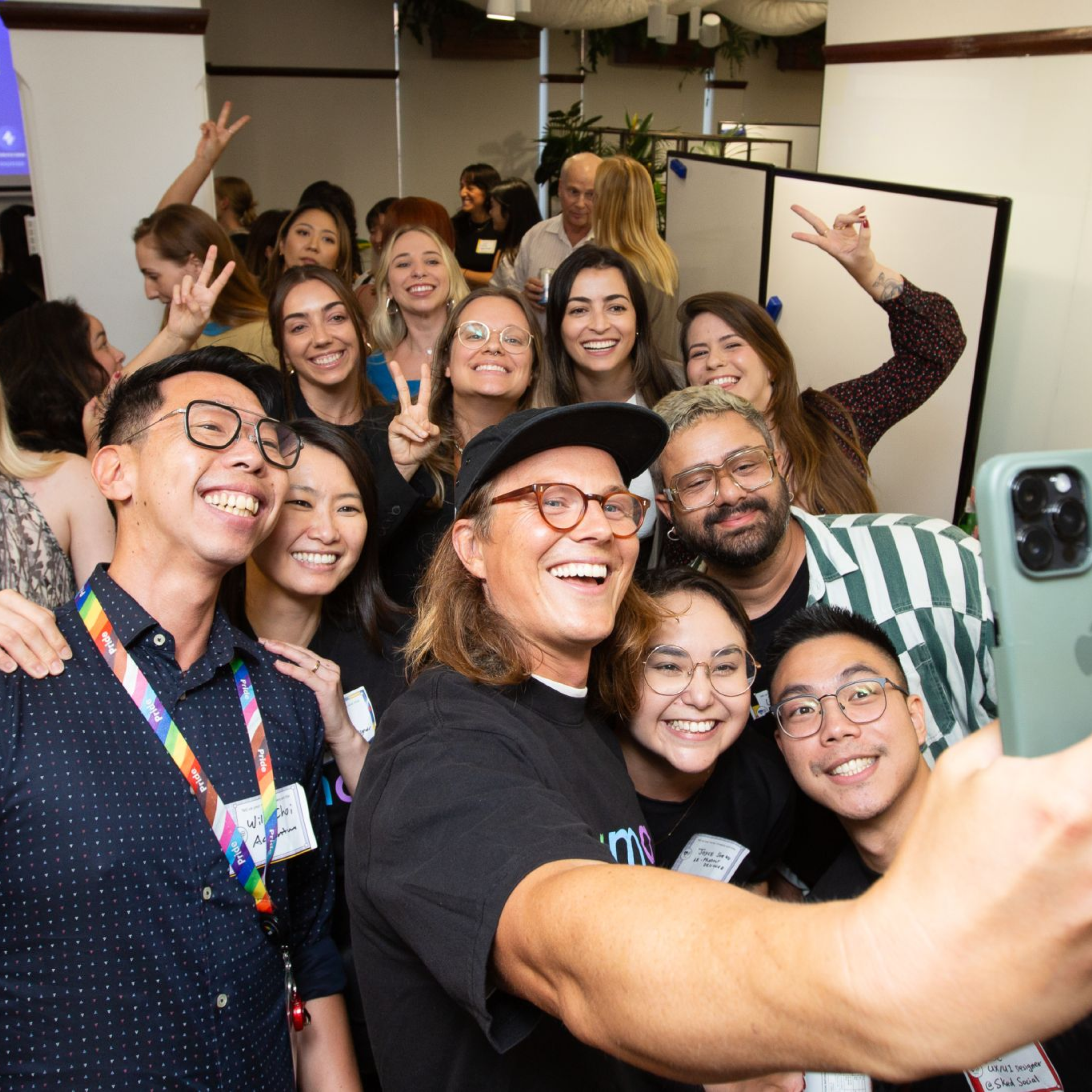 A group of people posing for a selfie at a lively event. They are smiling, laughing, and making peace signs, with a mix of colorful badges and lanyards visible. The room is bustling with energy and camaraderie.