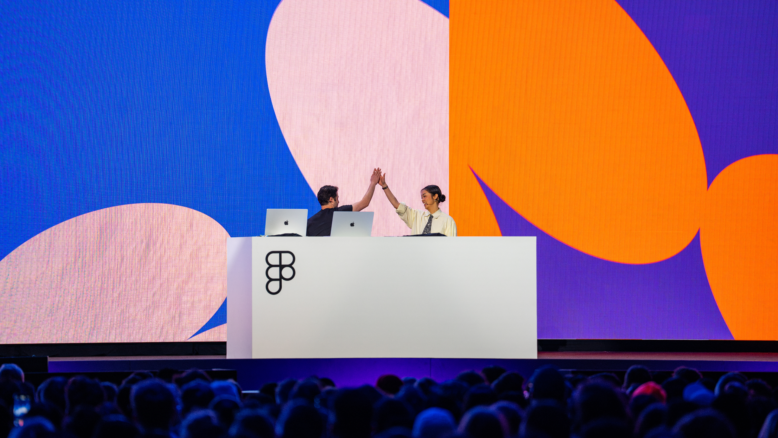 Two speakers on stage at Config high-five behind a white podium, standing in front of a colorful screen.