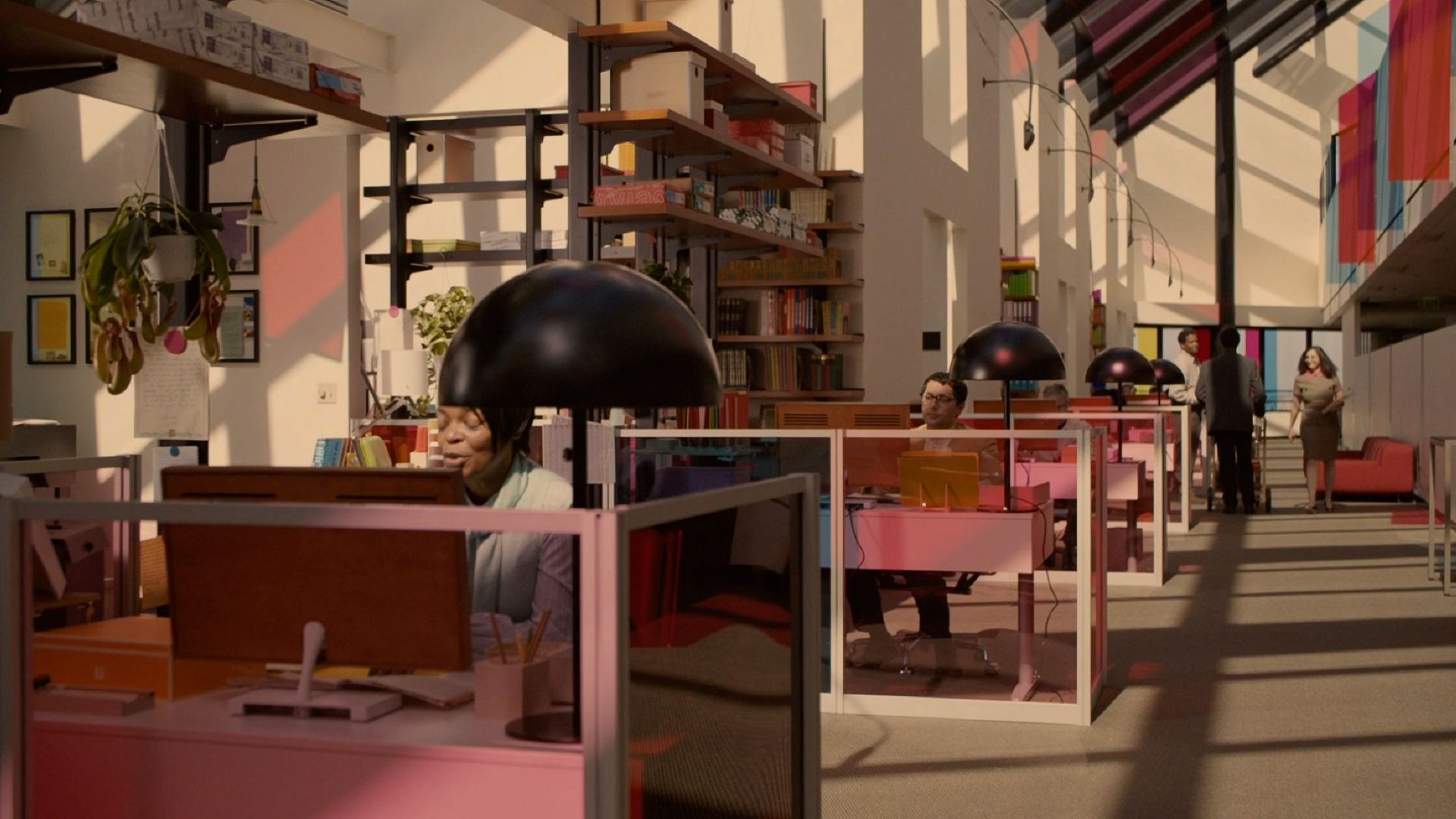 A bright, open-plan office with pink cubicles, bookshelves, and colorful glass panels glows in warm sunlight.