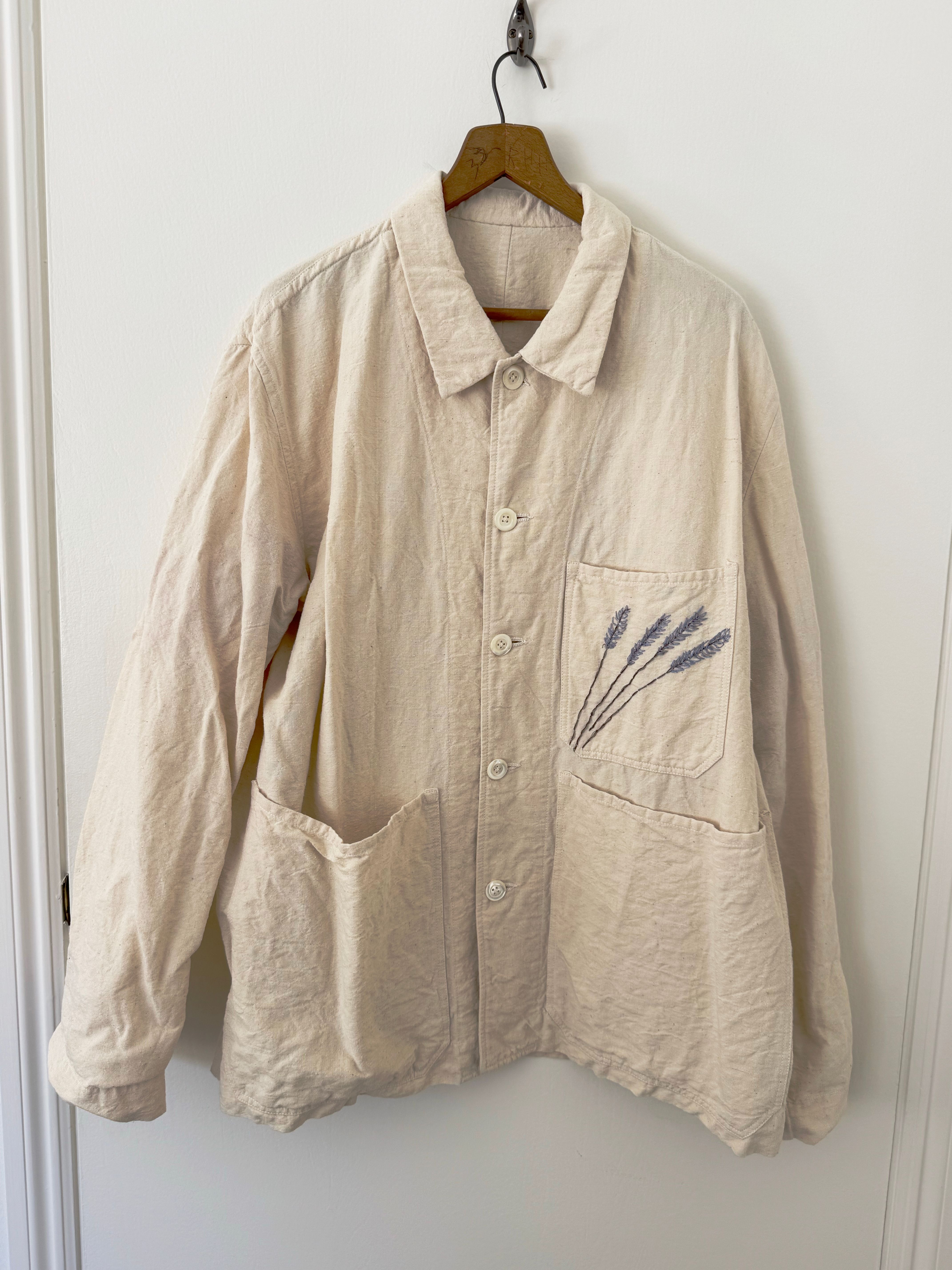Beige button-up jacket with a collar, featuring three front pockets and an embroidered lavender design on the upper left pocket, hanging on a wooden hanger against a white wall.