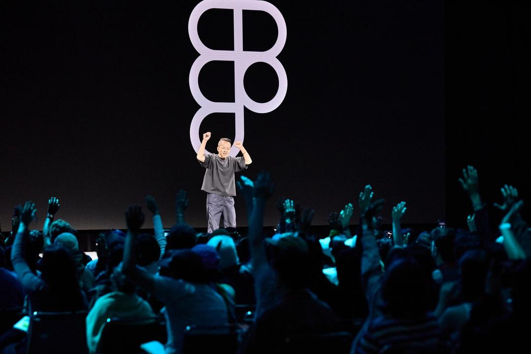 A photograph of a speaker on stage at a large conference, addressing an enthusiastic audience. The speaker, wearing dark casual clothing, stands with arms raised in front of a massive screen displaying a bold, abstract Figma logo. The audience, seated in rows, has many members raising their hands in engagement. The stage is dimly lit, with spotlights highlighting the speaker and the screen. The scene conveys excitement and energy, indicative of a keynote presentation or major announcement at a design-focused event.