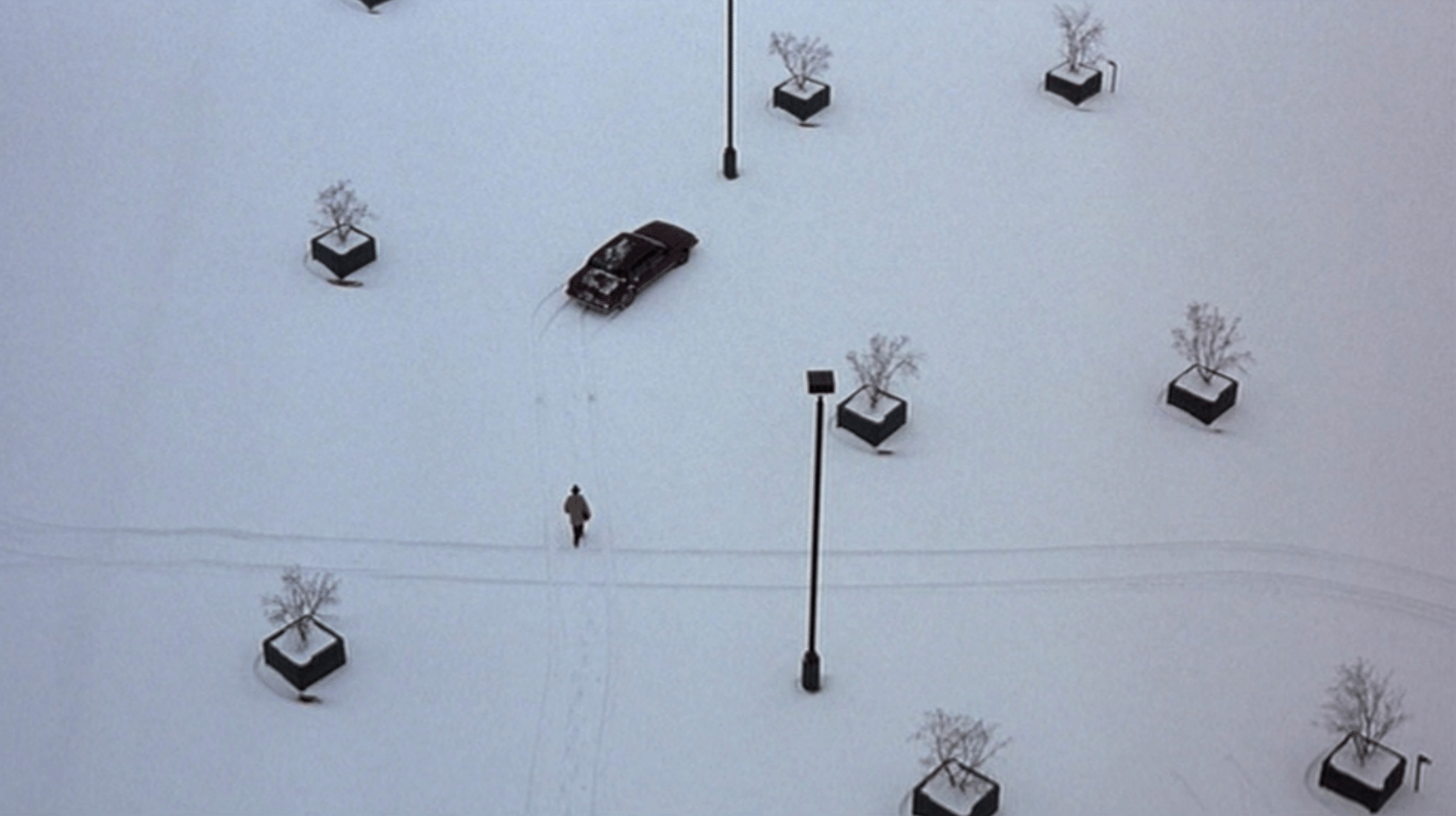 A lone figure walks across a snow-covered parking lot toward a car surrounded by evenly spaced planters and lampposts.