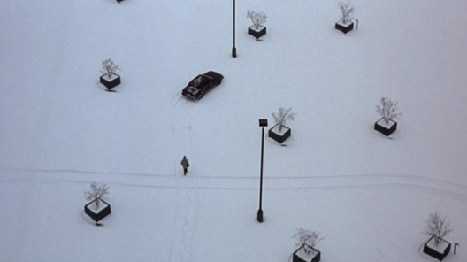 A lone figure walks across a snow-covered parking lot toward a car surrounded by evenly spaced planters and lampposts.