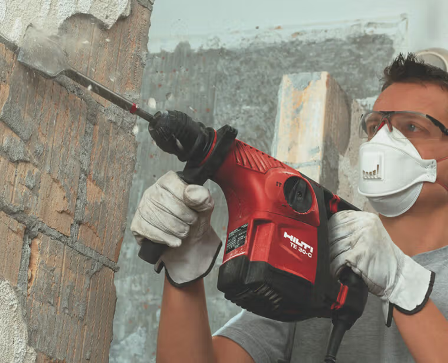 Chiselling plaster off a brick wall with a rotary hammer, wearing safety glasses, a dust mask and gloves