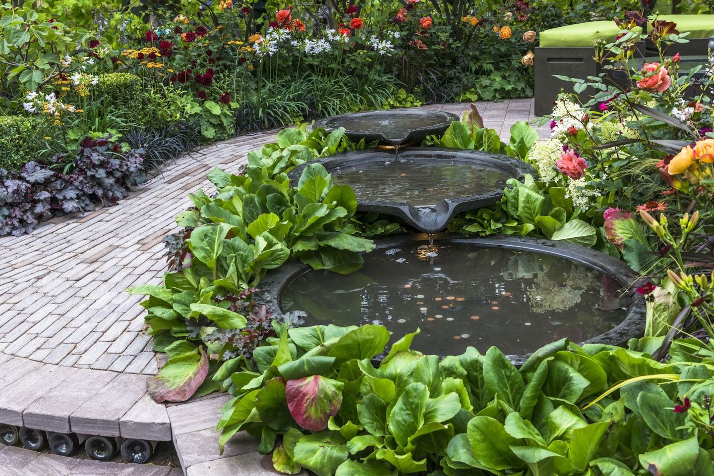 a triple level water feature fountain with small brick pavement in the background surrounded by lush garden plants