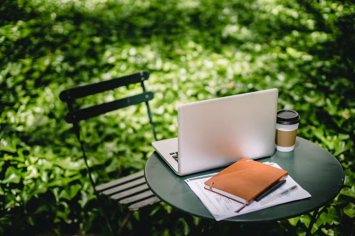 A chair and coffee table set up in the garden. The table has a laptop, notebook and a hot drink on top of it.