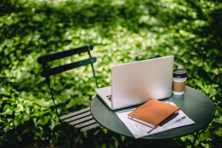 A chair and coffee table set up in the garden. The table has a laptop, notebook and a hot drink on top of it.