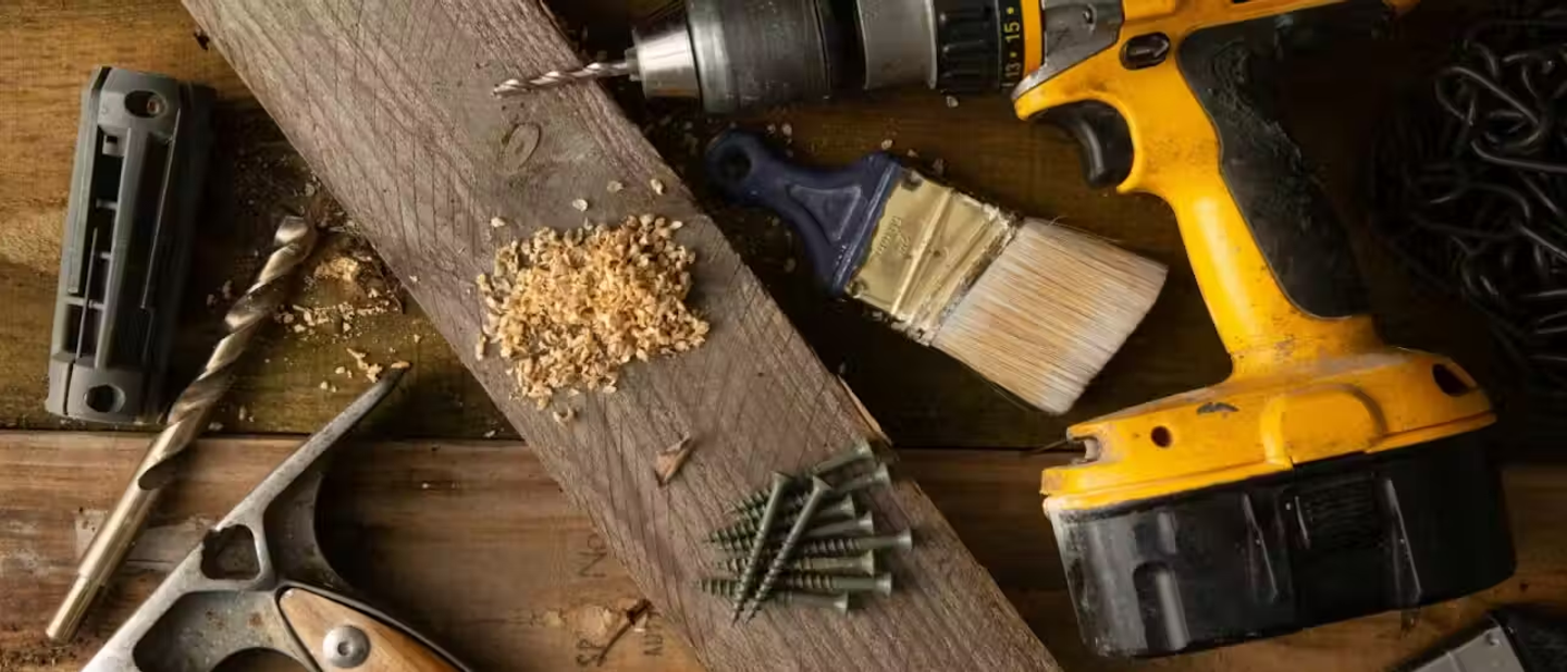 A table with a cordless drill, screws, a paintbrush and a plank of wood on top of it.