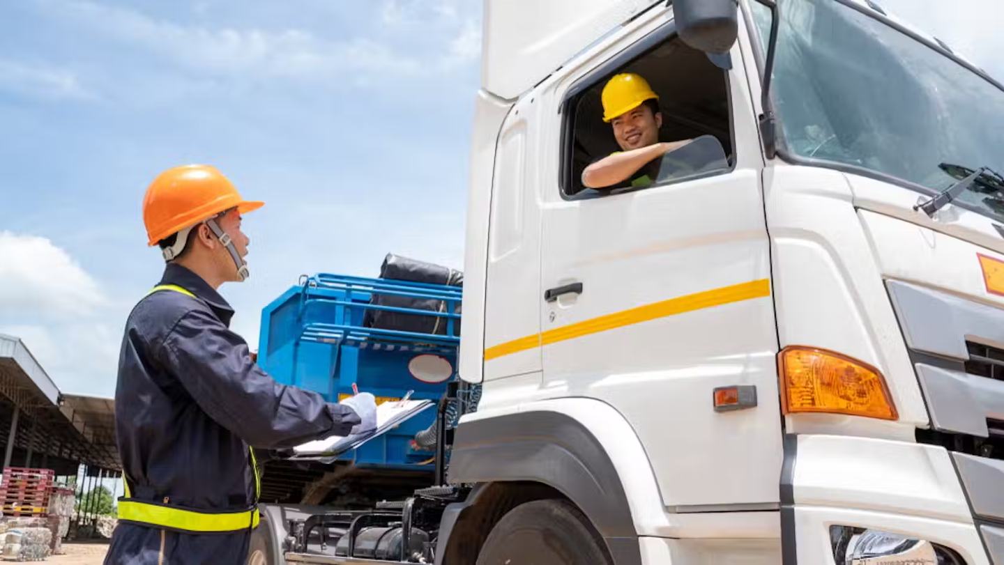 A construction worker with a clip board talks to a co-worker who is sat in the driver's seat of a lorry on site.