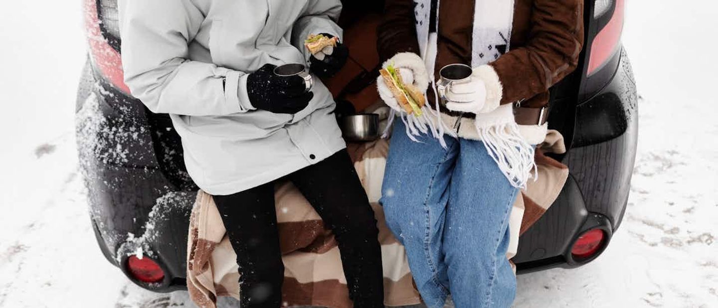 two people sitting in a trunk of a car eating sandwiches and drinking hot beverages