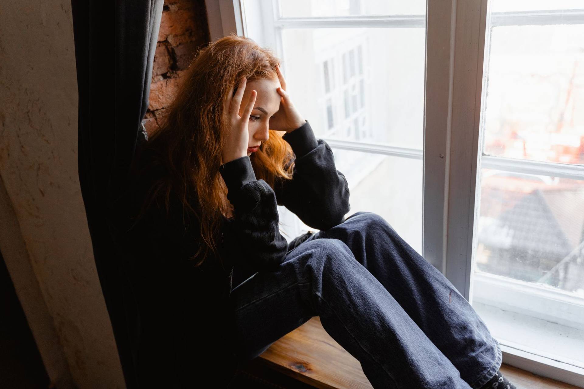 Une femme assise près d’une fenêtre, les mains sur le visage, l’air angoissé.